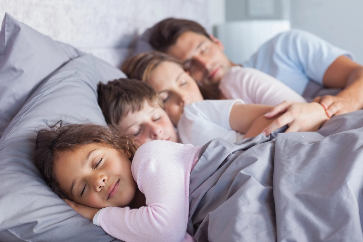 Schlafende Familie im Bett, zwei Erwachsene und zwei Kinder, grau-gestreifte Bettwäsche, gemütliche Atmosphäre.