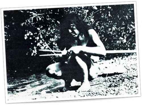 Ein Mann sitzt am Ufer eines Flusses und bearbeitet einen Speer. Er ist barfuß und trägt natürliche Kleidung.