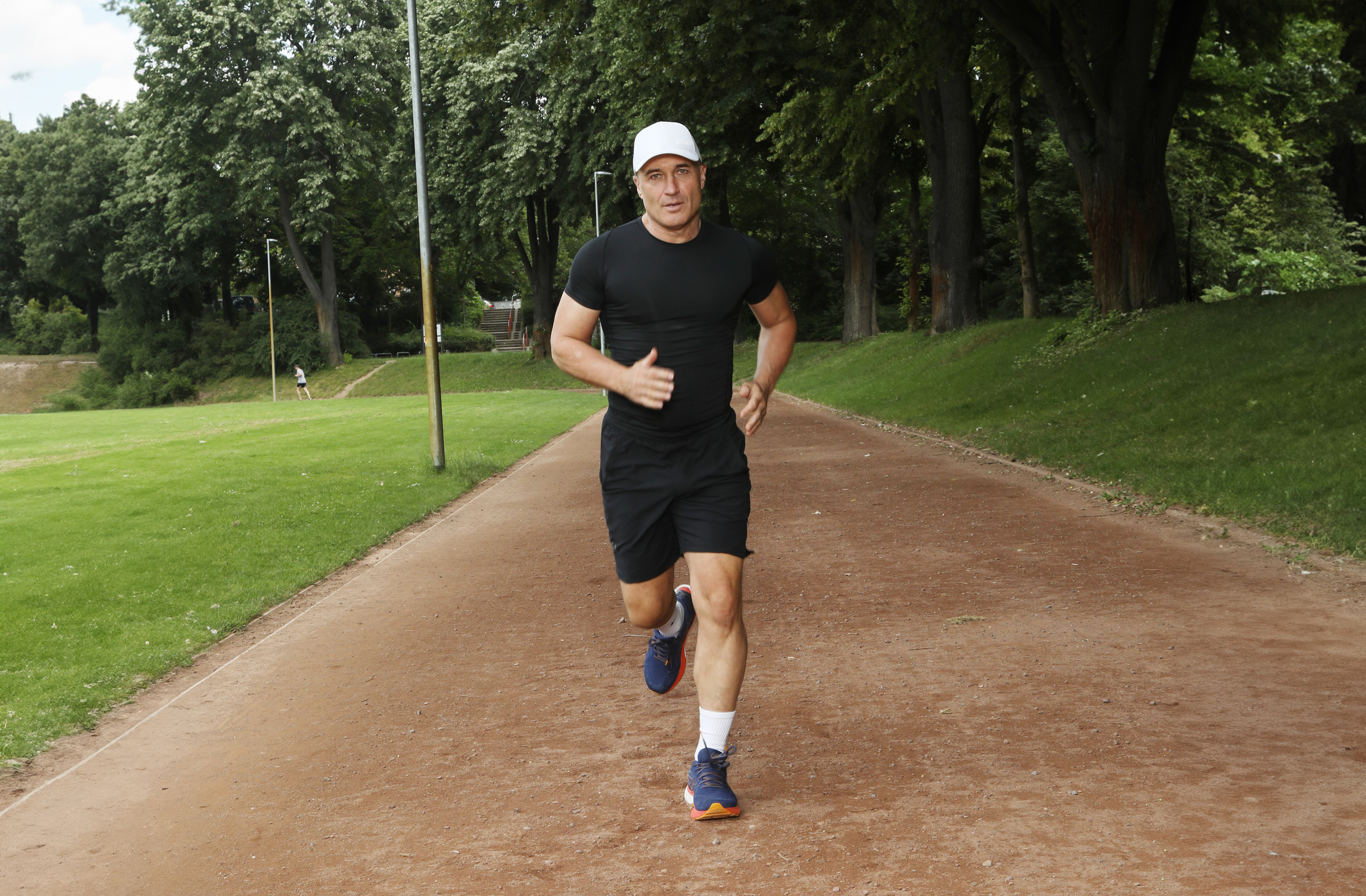 Laufschuhe Herren – Sportliche 10-km-Modelle Mann joggt auf einem Waldweg, trägt schwarze Sportkleidung und blaue Laufschuhe.