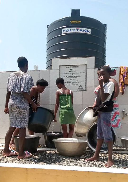Gruppe von Kindern entnimmt Wasser aus einem Wassertank, verschiedene Behälter, helle Kleidung, sonnige Umgebung.