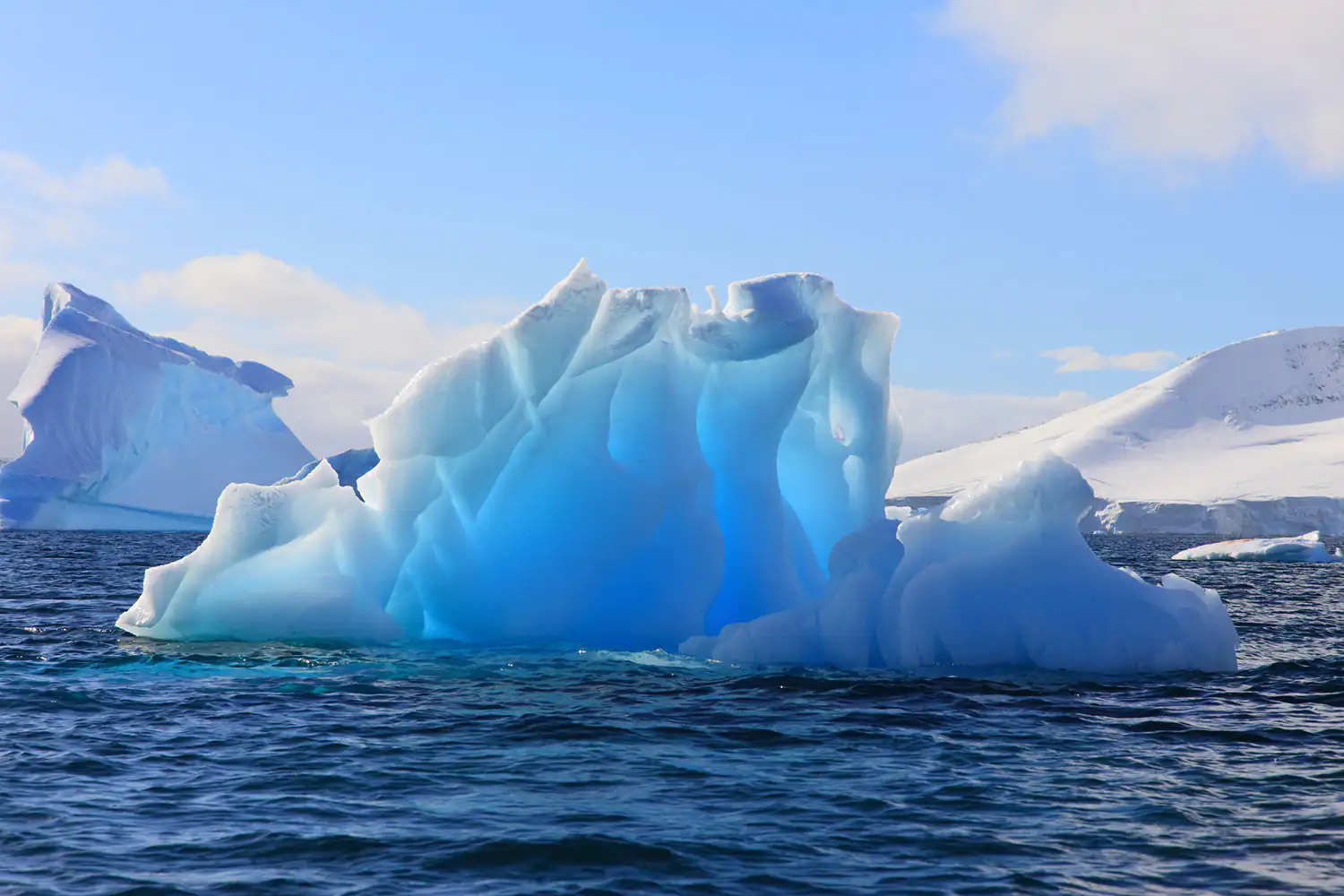 Ein blauer Eisberg schwebt im ruhigen Wasser, Umgebung zeigt schneebedeckte Berge und Himmel.