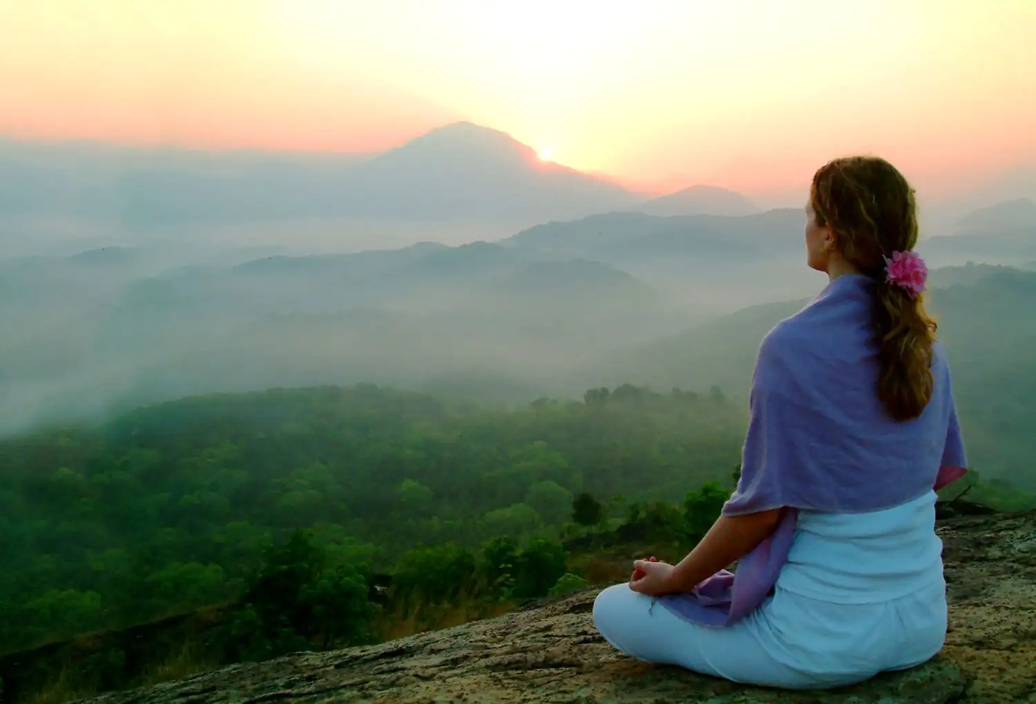 Frau sitzt meditierend auf einem Felsen, umgeben von einer nebligen Berglandschaft bei Sonnenaufgang.