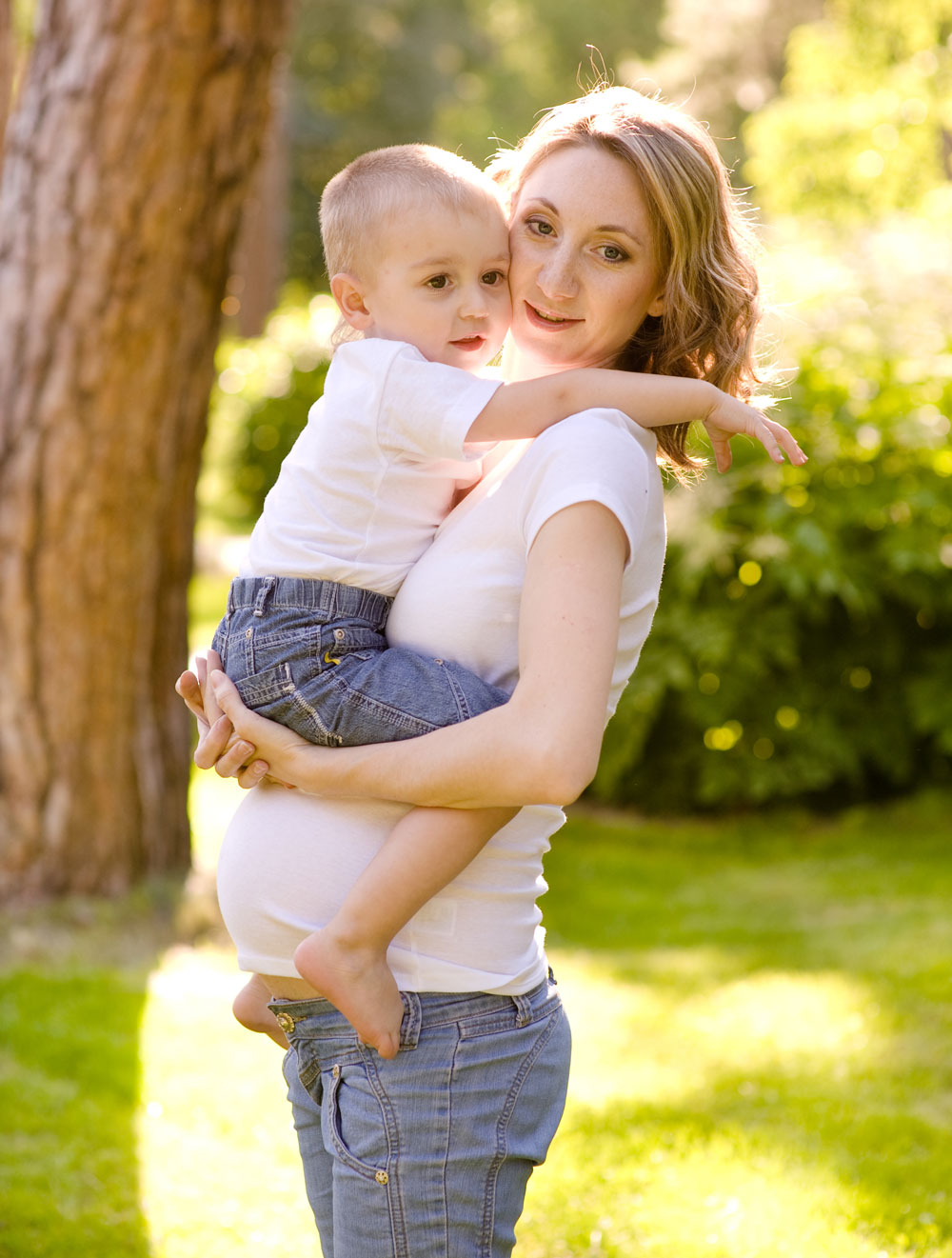 Eine schwangere Frau trägt ein Kind im Arm, im Freien, in einer grünen Umgebung.