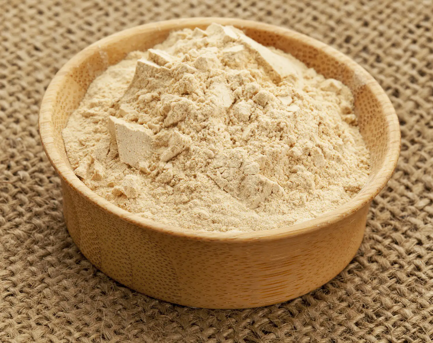 Fein gemahlenes Maca Pulver in einer Holzschale, beige Farbe, auf einer Jute-Oberfläche.